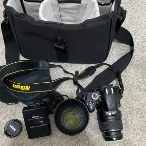kamra Nikon d3200  - Bilderna visar en Nikon D3200 systemkamera med två objektiv (18-55mm och 55-200mm), batteriladdare, kameraväska och tillbehör. Perfekt för dig som vill ta din fotografering till nästa nivå, oavsett om du är nybörjare eller vill utvecklas kreativt. Allt du behöver för att komma igång direkt!