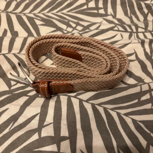 Beige flätat bälte med skinn-detaljer helt ny - Snyggt beige flätat bälte med bruna skinn-detaljer och silverfärgat spänne. Bältet har en stilren och klassisk look med vävd struktur som ger extra karaktär. Perfekt för att lyfta vilken outfit som helst med en touch av stil.