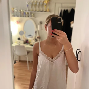 Vit linnetopp med spetskant - Säljer en vit linnetopp med tunna axelband och snygg spetskant vid urringningen. Toppen har en lös passform och är tillverkad i ett lätt, glansigt material som ger en fräsch och somrig känsla. Perfekt att styla med jeans eller kjol för en enkel look.