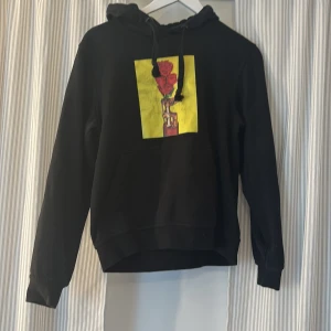 Svart hoodie med ros-tryck från Clique - Svart hoodie från Clique i storlek S med en röd ros i en brännare på en gul bakgrund. Tröjan har huva med snörning och ficka i fram. Inga större tecken på användning trycket är lite slitet. 