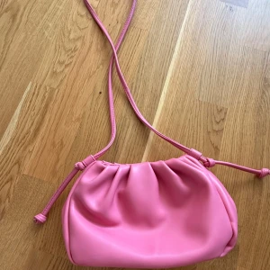 Rosa axelväska med knytdetaljer - Söt rosa axelväska i mjukt syntetmaterial med långa smala axelband och knytdetaljer på sidorna. Väskan har en puffig form och rynkade detaljer upptill som ger en trendig look. Perfekt för att lyfta vilken outfit som helst.