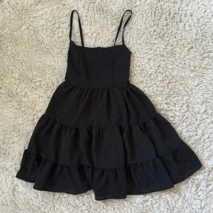 Svart kortklänning med tunna axelband - Supersöt svart kortklänning med tunna axelband och volangig kjol. Klänningen har diskreta ränder och smock i ryggen för extra komfort. Perfekt för dig som gillar en luftig och lekfull stil med lite edge.
