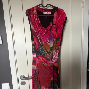 Färgstark Desigual klänning med mönster - Säljer en unik och färgstark klänning från Desigual med blommigt och abstrakt mönster i rött, rosa, orange och svart. Klänningen har draperad halsringning, är ärmlös och har en snygg knutdetalj framtill. Perfekt för dig som gillar att sticka ut med din stil!