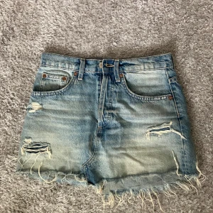 Blå jeanskjol med slitningar från Zara - Säljer en blå midwaist jeanskjol från Zara i modell kortkjol. Kjolen har coola slitningar och fransig nederkant för en avslappnad vibe. Klassisk femficksdesign och bälteshällor. Perfekt för dig som gillar en edgy och trendig stil.