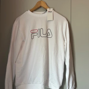 Vit sweatshirt från FILA med logga - Vit sweatshirt från FILA med stor logga i svart och rött framtill. Tröjan har rund halsringning, långa ärmar och är tillverkad i mjuk bomull. Perfekt för en clean och sportig look. Hör gärna av er vid frågor!