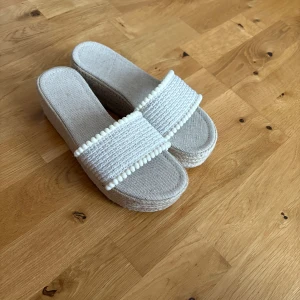 Beiga sandaler med platåsula - Snygga beiga sandaler med bred flätad rem och dekorativa små bollar. Platåsulan är klädd i tyg och ger extra höjd. Perfekta för varma dagar och ger en chill sommarkänsla till din outfit.