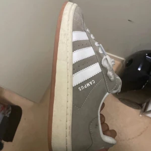 Adidas Campus grå sneakers mocka - Säljer ett par Adidas Campus sneakers i grå mocka med klassiska vita ränder och vit sula. Skorna har rund tå, platt gummisula och snörning. Perfekta för dig som gillar en clean och tidlös streetstil.