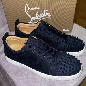 Svarta sneakers med nitar från Christian Louboutin - Säljer ett par marinblå sneakers från Christian Louboutin med marinblå nitar på tån och klassisk röd sula. Skorna har snörning, rund tå och är gjorda i mocka med vita platta sulor. Perfekta för dig som vill sticka ut med en lyxig touch. Har låda och allt Kom med byten 