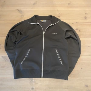 Grå zip-jacka från Carhartt - Säljer en svart zip-jacka från Carhartt med vit dragkedja och vita detaljer vid fickorna. Jackan har ribbade muddar och krage samt två snedställda fickor framtill. Den har en liten skada på ena mudden men den är lagad och märks knappt. Skicka ett meddelande för frågor, storlek, mått osv. 