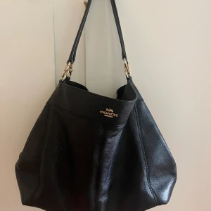 Svart axelväska från Coach - Säljer en svart axelväska från Coach i äkta skinn med guldfärgade metalldetaljer och klassisk logotyp framtill. Väskan har en rymlig form och bred axelrem, perfekt för att bära alla dina grejer med stil.
