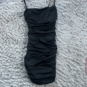 Svart tight kortklänning med rynkade detaljer - Säljer en svart tight kortklänning med smala axelband och snygga rynkade detaljer längs hela klänningen. Perfekt för dig som vill ha en enkel men ändå stilren look. Klänningen har en figurnära passform och är superenkel att styla. Storlek xs men kan passa större, vet ej vart ifrån