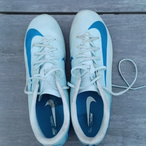 Nike Mercurial fotbollsskor i vitt och blått - Säljer ett par Nike Mercurial fotbollsskor i vitt med blå detaljer och snörning. Skorna har rund tå, platt sula med dobbar och är perfekta för dig som vill ha stilrena och snabba skor på planen.  Det tillkommer även skillsocks i storlek L och skokartong. Pris kan diskuteras. Strumporna åker med om man vill ha dom men man kan välja att strunta i de. Strumporna gör inte paket dyrare vill bara bli av med dom.