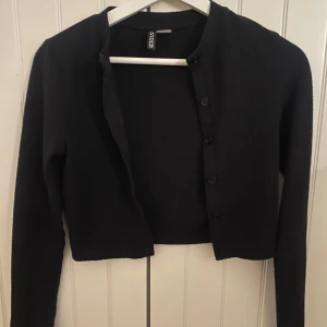 Svart croppad kofta från H&M Divided - Säljer en svart croppad kofta från H&M Divided. Koftan har långa ärmar och knäppning framtill med svarta knappar. Enkel och stilren modell som passar perfekt till många olika outfits.