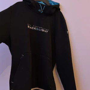 Svart hoodie från Peak Performance Black Light - Svart hoodie från Peak Performance Black Light-kollektionen med turkosblått innerfoder i huvan och broderad logga på bröstet. Praktisk magficka framtill och dragsko i huvan. Perfekt för en sportig och avslappnad stil.