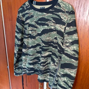 Grön camouflage tröja från Replay - Säljer en långärmad tröja från Replay med grön och svart camouflage-mönster. Tröjan har rund halsringning och en dragkedjedetalj nertill på båda sidor. Perfekt för dig som gillar streetwear och vill sticka ut lite extra. Pris kan diskuteras vid snabb affär