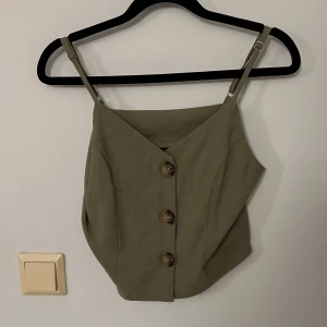 Grön linnetopp med knappar - Snygg grön linnetopp med justerbara axelband och stora dekorativa knappar framtill. Toppen har en v-ringad skärning och är tillverkad i ett mjukt, lätt material. Perfekt för varma dagar och enkel att matcha med olika outfits.