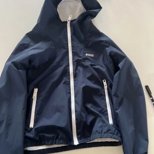 Mörkblå vindjacka med huva från BOSS - Snygg mörkblå vindjacka från BOSS med vit dragkedja och huva. Jackan har två fickor med dragkedja och meshfoder på insidan för extra komfort. Endast använd fåtal gånger! Den är i storleken 14-s (barnstorlekar) vilket motsvarar ungefär s-xs i (herrstorlekar). Ny pris är ca 2000 kr och mitt pris är 379kr. Perfekt för blåsiga dagar och enkel att matcha med olika outfits. Skriv bara vid minsta fråga, priset kan diskuteras! 