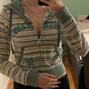 Randig hoodie från urban outfiters  - Säljer en randig hoodie i grönt, vitt och lila färg med dragkedja och huva. Tröjan har broderad text och siffran 98 framtill samt ribbade muddar och fickor. Perfekt för en avslappnad stil.