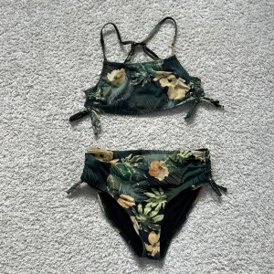 Grön blommig bikini med knyt - Snygg bikini med tropiskt blommönster i grönt och gult. Överdelen har smala axelband och knyt i sidorna, och bikinitrosan har hög midja och knyt i sidorna. Perfekt för strandhäng eller poolen!