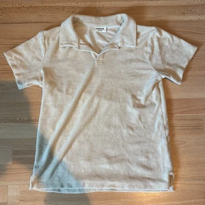 Beige pikétröja från Kappahl - Säljer en beige pikétröja från Kappahl i frottéliknande material. Tröjan har korta ärmar och klassisk krage med liten v-ringning. Perfekt för en avslappnad och stilren look.