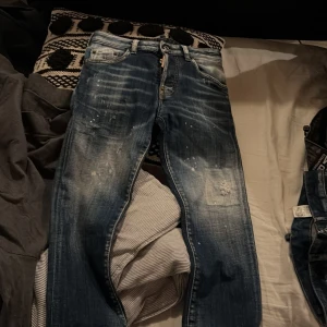 Dsquared2 jeans med slitningar och tryck - Säljer ett par blå Dsquared2 jeans med tydliga slitningar och ljusa partier framtill. Byxorna har klassisk femficksmodell, knappgylf och Dsquared2-detaljer på etikett och innerfoder. Snyggt tryck med siffran 64 på bakfickan.