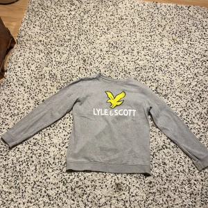 Grå sweatshirt från Lyle & Scott - Grå sweatshirt från Lyle & Scott med klassisk gul logga och vit text på bröstet. Tröjan har rund halsringning och långa ärmar. Perfekt för en avslappnad och stilren look.
