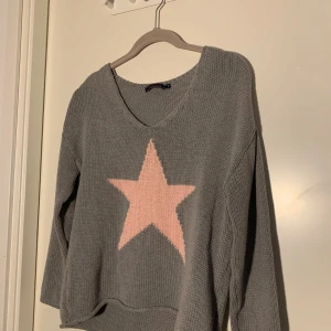 Grå stickad tröja - Supermjuk grå stickad tröja från Bondelid med en stor rosa stjärna framtill. Tröjan har v-ringning och långa ärmar, perfekt för lager-på-lager. Broderad Bondelid-logga nertill. Snygg och enkel att matcha!