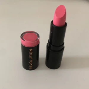 Rosa läppstift från Revolution - Säljer ett snyggt nytt rosa läppstift från Revolution. Hylsan är svart med guldtext och har en rosa detalj i botten som matchar färgen på läppstiftet. Perfekt för dig som vill ha en poppig färg på läpparna.
