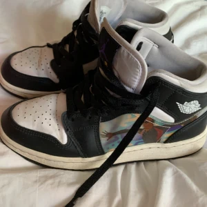 Nike Air Jordans svartvita sneakers - Säljer ett par klassiska svartvita Nike Air Jordans med högt skaft och svart snörning. Skorna har vit tå och sula, svart ovandel och detaljer samt en färgglad illustration på sidan. Perfekta för dig som gillar streetwear och sneakers. Skorna är storlek 36. Ingen låda som hör till men jag har en helt random Nike låda som du får!