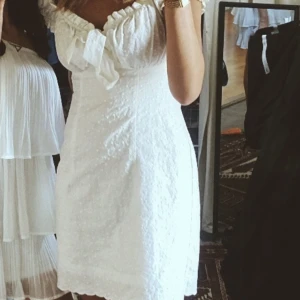 Vit kortklänning med volang och rosett - Superfin vit kortklänning med volangkant upptill och söt rosettdetalj framtill. Klänningen har offshoulder-modell och är figurnära med lätt struktur i tyget. Perfekt för sommarens alla tillfällen! Köpt för 950kr❤️