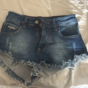 Jeansshorts från Tommy Hilfiger - Säljer ett par blå jeansshorts från Tommy Hilfiger med fransig kant, klassisk femficksdesign. Shortsen är midwaisted, (ser högmidjade ut på mig då jag har kort överkropp)Perfekta för varma dagar och enkel att matcha. Midjemått 34,5cm men tyget är stretchigt