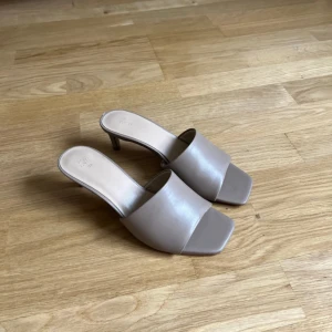 Beige slip-in klackskor med fyrkantig tå - Säljer ett par stilrena beige slip-in klackskor med öppen häl och fyrkantig tå. Skorna har en låg klack och bred rem över foten, vilket ger en modern och minimalistisk look. Perfekta för dig som gillar enkel och trendig stil. Jag köpte dom begagnade men aldrig använt dom själv!