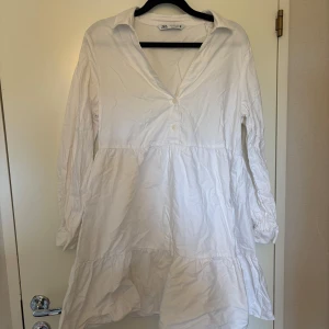Vit skjortklänning från Zara - Säljer en vit skjortklänning från Zara med v-ringning och krage. Klänningen har lång ärm och volang nertill, perfekt för en avslappnad och stilren look.