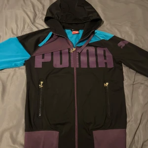Svart, blå och lila hoodie från Puma - Säljer en snygg hoodie från Puma med stor logga framtill. Jackan är svart och  detaljer på axlar och ärmar, samt lila muddar och dragkedja. Två fickor med dragkedja och huva. Perfekt för en sportig look.