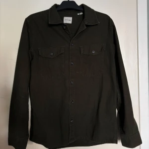 Mörkgrön overshirt från Jack & Jones - Säljer en mörkgrön overshirt från Jack & Jones med två bröstfickor och knäppning framtill. Skjortan har klassisk krage och långa ärmar, perfekt att bära som lager över en t-shirt.                                                                                
