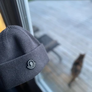 Grå mössa från Moncler - Säljer en stilren grå ribbstickad mössa från Moncler med uppvikt kant och klassisk logotyp framtill. Perfekt accessoar för kyliga dagar och enkel att matcha med olika outfits.