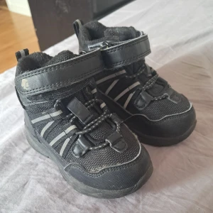 Svarta sneakers med kardborreband och reflexdetaljer - Säljer ett par svarta sneakers med praktiska kardborreband och elastiska snören. Skorna har reflexdetaljer på sidorna och är tillverkade i syntetmaterial med meshpartier för extra andningsförmåga. Perfekta för vardagsbruk.