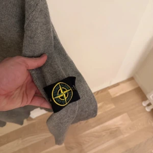 Grå stickad tröja från Stone Island - Säljer en grå stickad tröja från Stone Island med klassisk rund halsringning och ribbade muddar. Tröjan har den ikoniska Stone Island-loggan på ärmen och är perfekt för dig som gillar stilrena och exklusiva plagg. Mindre i storlek.