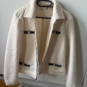 Vit teddyjacka från Holly & Whyte - Säljer en vit teddyjacka från Holly & Whyte med dekorativa pärlknappar och svarta detaljer vid fickorna och längs öppningen. Jackan har klassisk krage och lång ärm, perfekt för en trendig look.