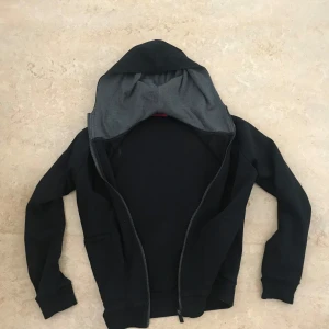 Svart hoodie från Hugo Boss - Säljer en svart hoodie från Hugo Boss med dragkedja och grå insida på huvan. Klassisk modell med långa ärmar och ribbade muddar. Perfekt för en avslappnad stil. Storlek S. 