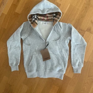 Grå burberry zip/kofta - Snygg grå hoodie från Burberry med klassiskt rutigt foder i huvan och dragkedja framtill. Tröjan har långa ärmar, två fickor och ribbade muddar. Perfekt för en avslappnad och stilren look. Helt ny med etikett. Storlek S men passar även XS nog. 