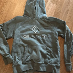 Olivgrön hoodie från GANT - Säljer en olivgrön hoodie från GANT med stor broderad logga på bröstet. Tröjan har huva, magficka och ribbade muddar vid ärmslut och nederkant. 