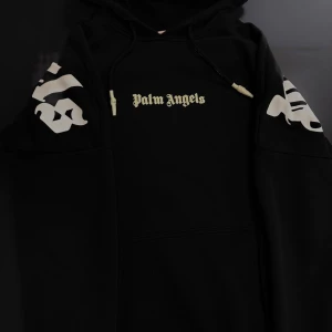 Svart hoodie från Palm Angels - Säljer en svart hoodie från Palm Angels med vit logga på bröstet, ärmarna och stor text på ryggen. Tröjan har huva med dragsko och en stor magficka. Tillverkad i mjuk bomull, perfekt för en avslappnad streetwear-look.