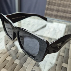Svarta solglasögon från Prada - Stilrena svarta solglasögon copia one från Prada med breda skalmar och markerad rektangulär form. Glasögonen har mörka linser och diskret Prada-logga på sidan ....mvh