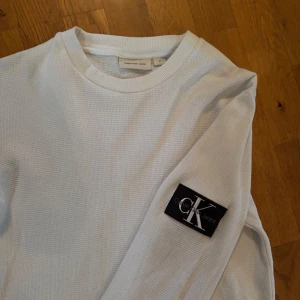 Vit sweatshirt Calvin Klein - Säljer en vit långärmad tröja från Calvin Klein Jeans. Tröjan har en svart Calvin Klein logga på ena ärmen. Klassisk rund halsringning och mjukt tyg.