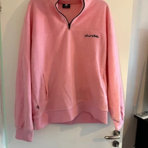Rosa fleece-tröja - Mysig rosa fleece-tröja med halv dragkedja och broderad logga på bröstet. Tröjan har långa ärmar, fickor på sidorna och ribbade muddar vid ärmslut och nederkant. Perfekt för kyliga dagar.