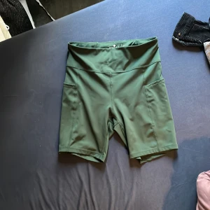 Gröna träningstights shorts från Ronhill - Mörkgröna träningstights shorts från Ronhill med hög midja och praktiska sidofickor. Tillverkade i stretchigt syntetmaterial som passar perfekt för löpning eller gym. Snygg och sportig design med diskret logga bak.
