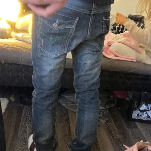 Mörkblå jeansbyxor med gröna detaljer - Säljer ett par mörkblå jeansbyxor från M2 med snygga slitningar och gröna sömdetaljer på bakfickorna. Klassisk femficksmodell med knapp och dragkedja. Perfekta för dig som gillar en stilren men ändå lite edgy look.