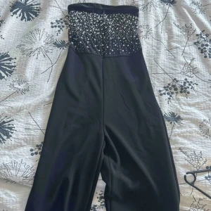 Svart byxdress med pärldetaljer -  svart byxdress med strapless överdel dekorerad med massor av pärlor i olika storlekar. Byxdressen har raka långa ben och en elegant, figurnära passform upptill.