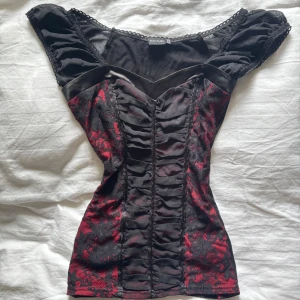 Svart och röd korsettopp med spetsdetaljer - Säljer en snygg svart korsettopp från Bodyflirt med röda spetsdetaljer och rynk framtill. Toppen har breda axelband och en figurnära passform som framhäver midjan. Perfekt för dig som vill sticka ut!